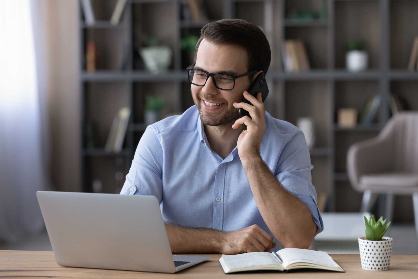 Happy young Caucasian businessman sit at desk at home office work online on computer talk with client or customer on cellphone. Smiling man use laptop gadget at workplace have smartphone conversation.